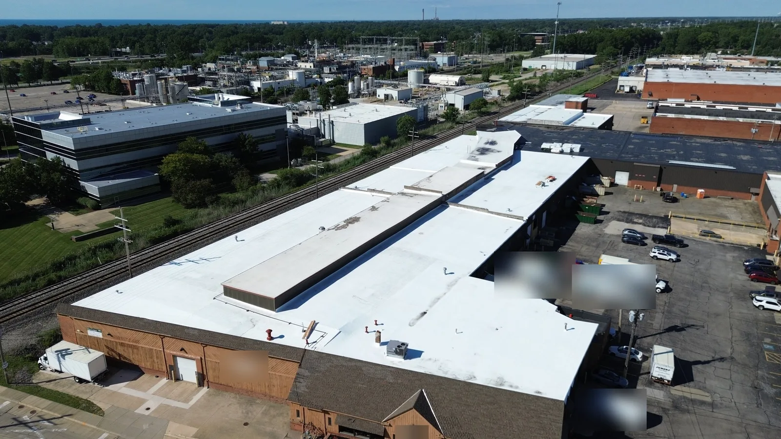 Drone view of a new white Carlisle TPO roof on a one-story industrial warehouse in Cleveland, Ohio. The photo shows raised center sections, rooftop units, a rail line alongside, and Lake Erie on the horizon.