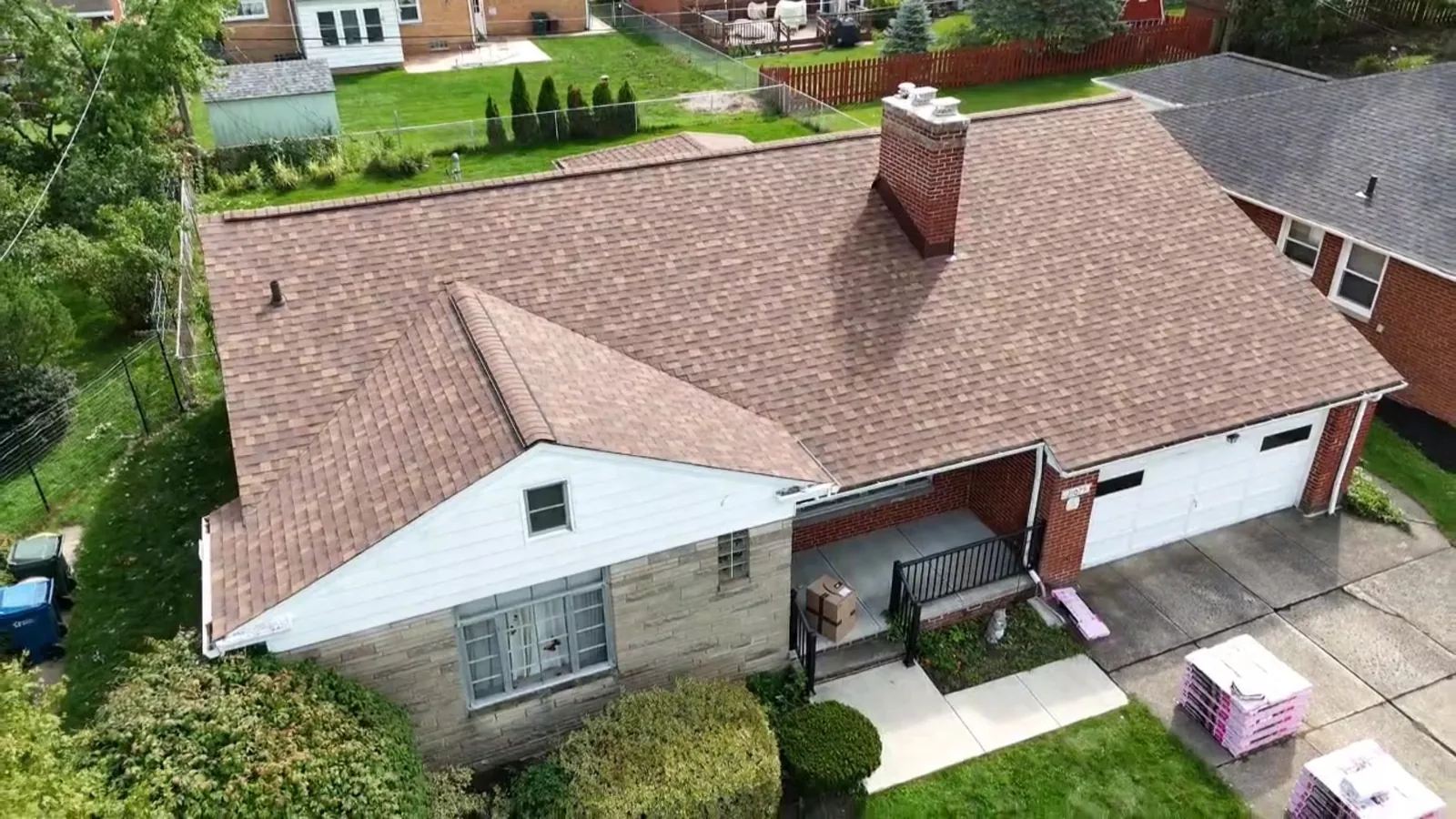 Aerial view of a brick and white-sided home with an attached garage in Fairview Park, Ohio, with a new Owens Corning Duration Desert Rose roof and shingle bundles in the driveway