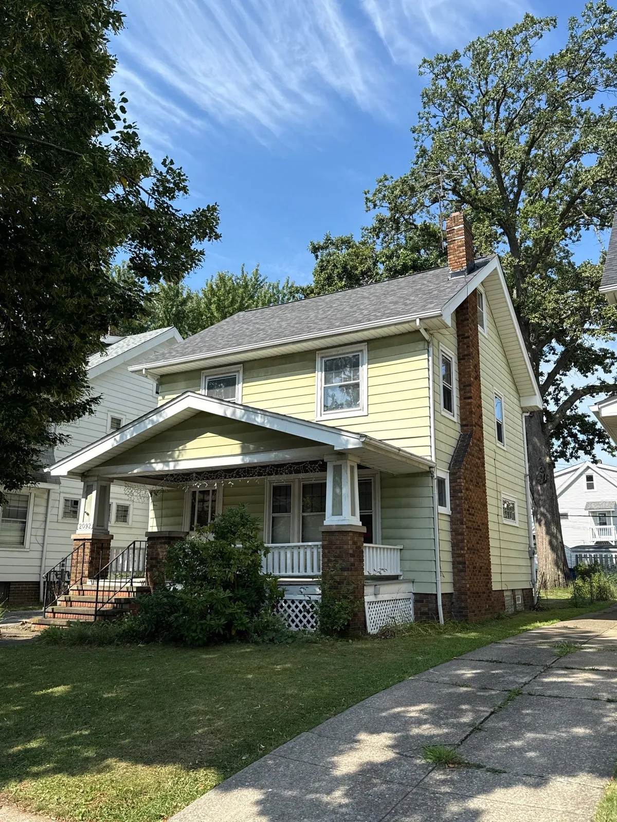 Yellow two-story home with a brick chimney, front porch, and new gray shingle roof under tall trees in Lakewood, Ohio