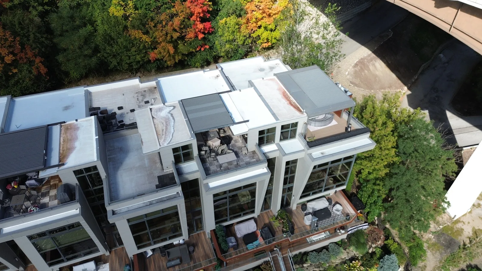 Drone view of a hillside townhome building in Rocky River, Ohio. It shows stepped flat roofs with new Carlisle TPO and silicone roof coating, rooftop terraces, fall foliage along the Rocky River valley, and a bridge in the upper corner.